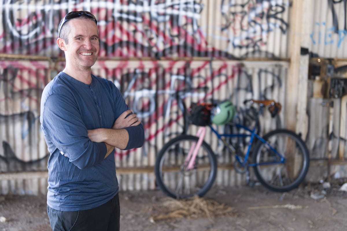 Simon stands in the foreground, with a slightly blurry bicycle in the background. The bike is leaning up against a grafittied corrugated steel wall. The ground is dirt.