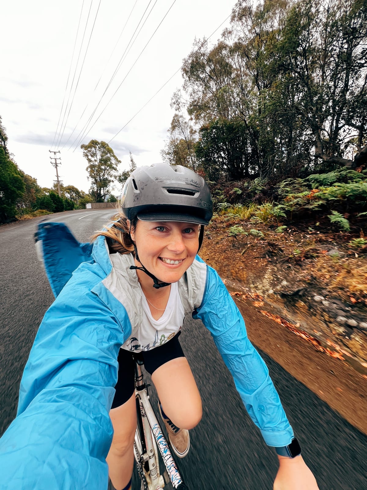 Caz, taking a selfie while riding a bicycle, wearing a blue rainjacket flapping in the wind, with a black helmet. The blitumen road is slightly blurred from the speed.